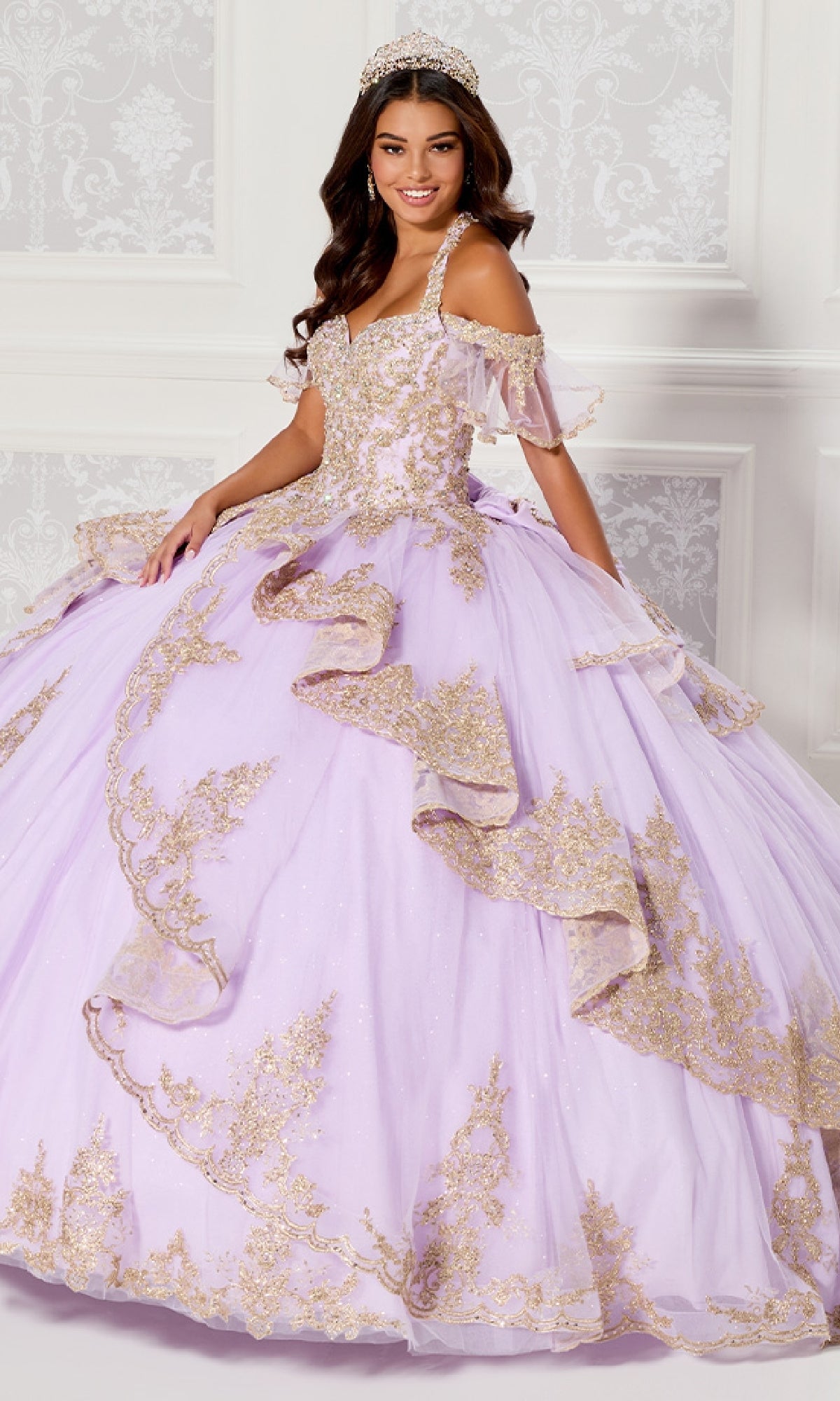 A woman smiles at the camera in a white, ornate room while wearing the Quinceanera Dress PR30118 By Princesa—a pastel lilac gown with gold glitter embroidery—paired with a sparkling tiara.